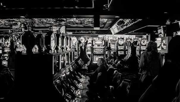 A dimly lit casino filled with rows of slot machines, some occupied by people. The machines' lights contrast with the dark surroundings, creating a vibrant yet subdued atmosphere.