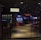 A dimly lit casino with several rows of slot machines, some displaying bright graphics and lights. There are a few people scattered throughout the area, engaging with the machines. The floor is reflective and polished, adding to the ambiance, while the ceiling is dotted with numerous small lights.