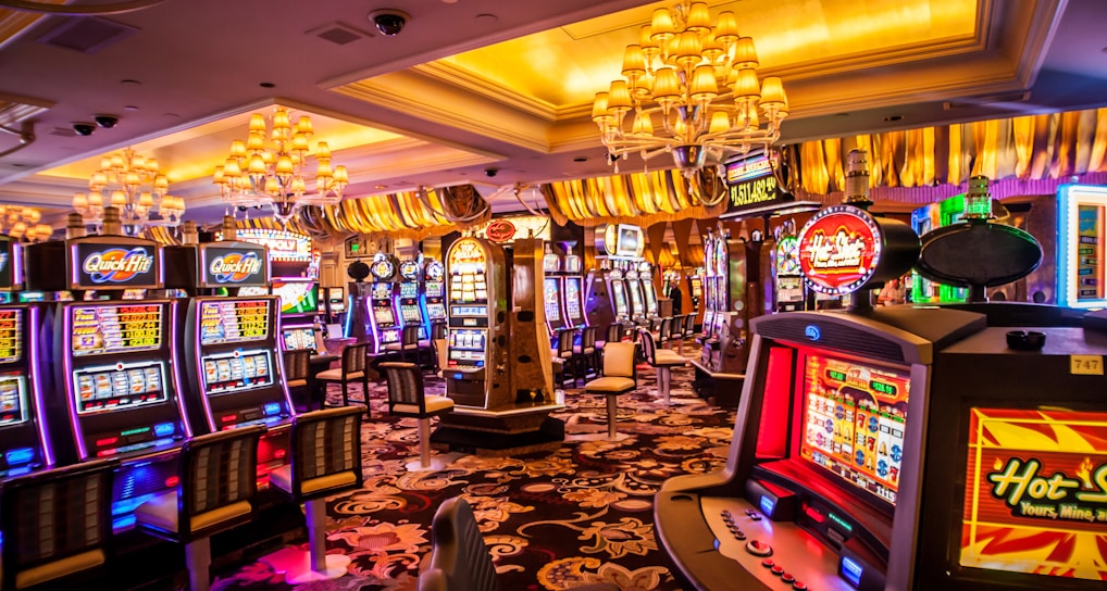 A vibrant casino interior filled with rows of colorful slot machines, each illuminated with bright lights and displays. The ceiling is adorned with elegant chandeliers and golden drapes, creating a luxurious atmosphere. The patterned carpet adds to the elaborate design of the space.